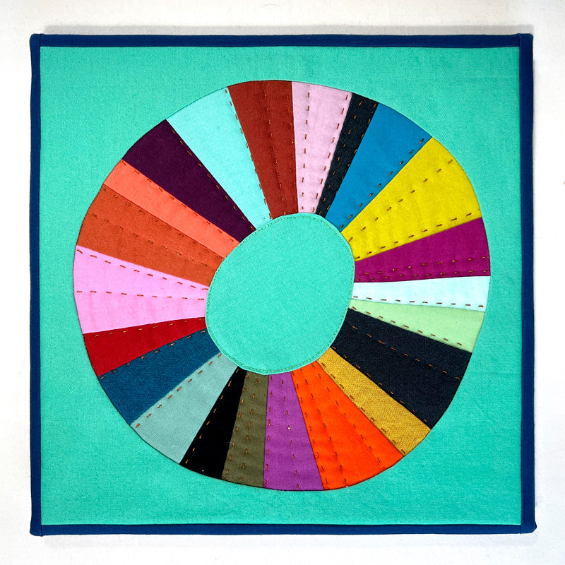 Small quilt art in bright jewel tone colors. The shape is a wonky circle on a green background with gold hand stitching.