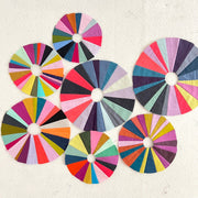 A wonky sewn circles on a cream background. The circle is composed of wedges in shades of pink, blue and orange.