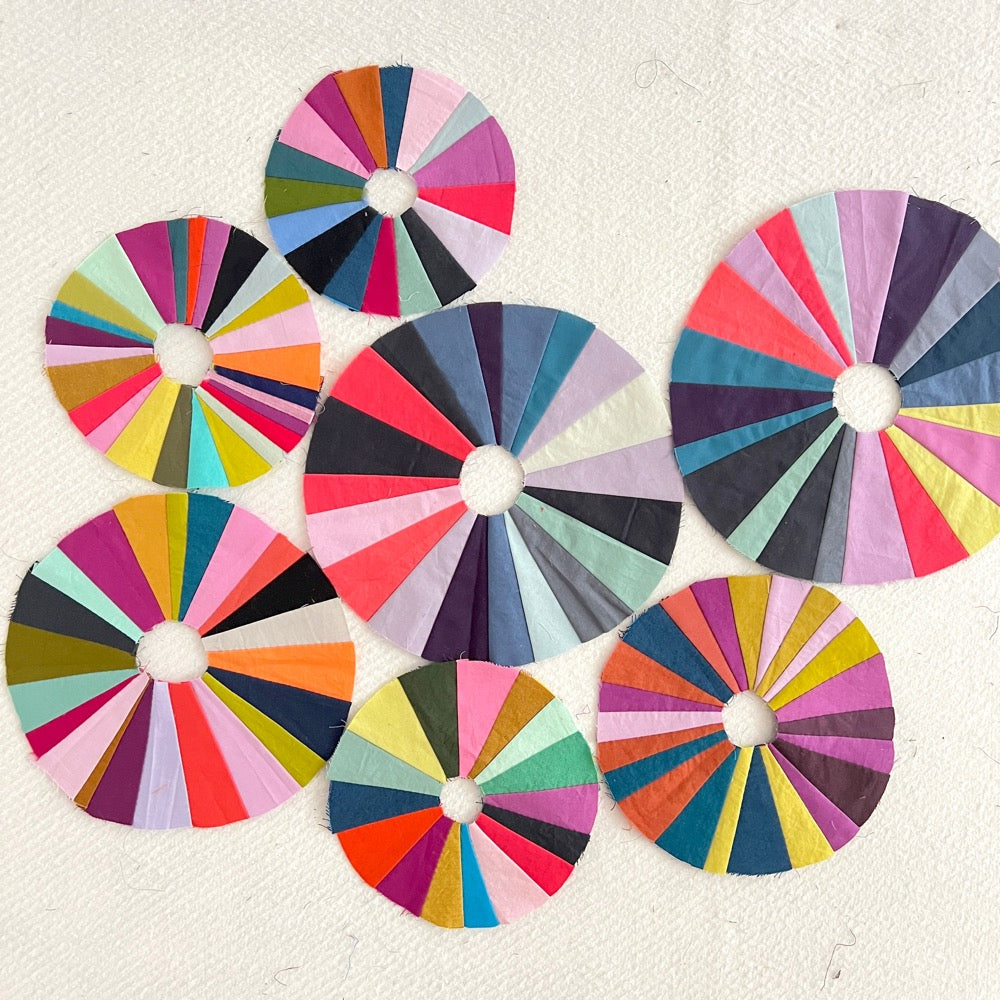 A wonky sewn circles on a cream background. The circle is composed of wedges in shades of pink, blue and orange.