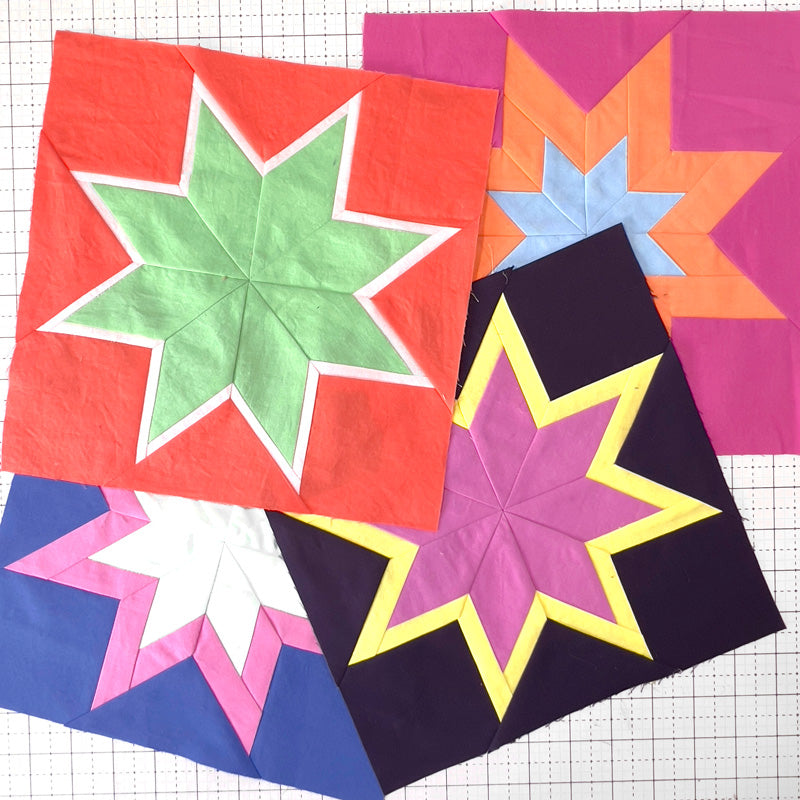 Four fabric star quilt blocks arranged on a white grid cutting mat. The blocks feature geometric star patterns in red, green, orange, blue, purple, pink, yellow, and black.