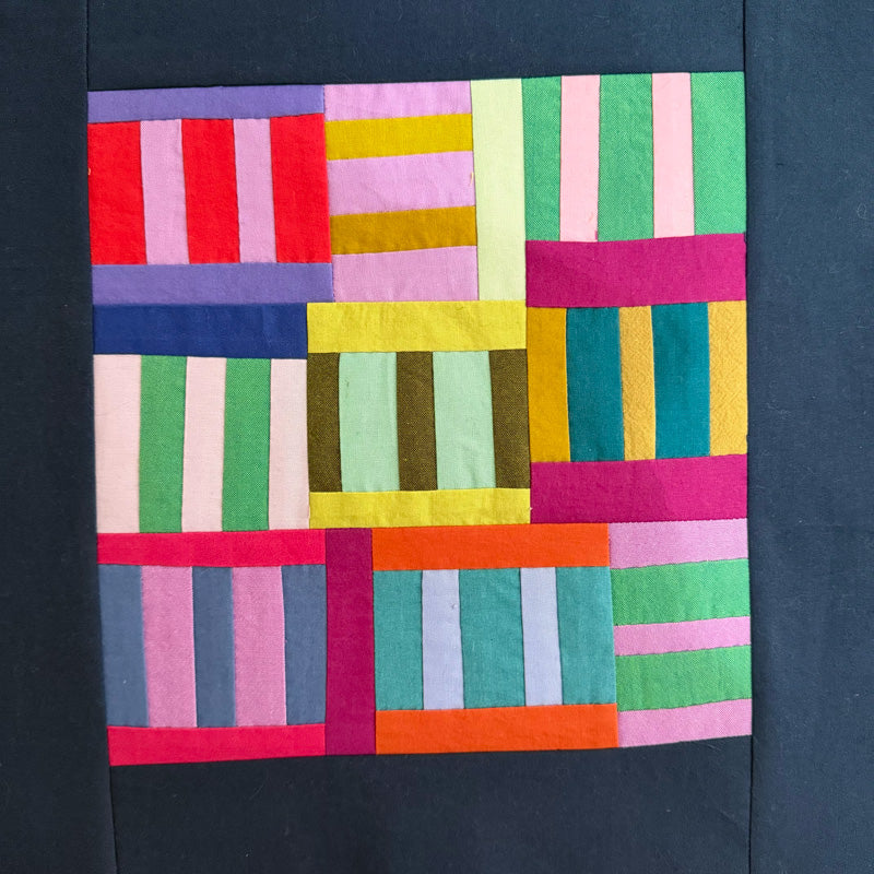 A colorful patchwork quilt or fabric art piece with vertical and horizontal striped blocks in pink, green, yellow, blue, orange, purple, and white, displayed against a dark navy fabric background.