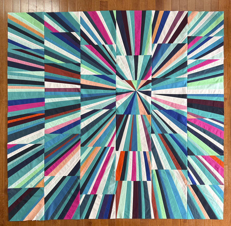 A square quilt featuring a vibrant geometric starburst pattern with radiating stripes in teal, blue, pink, orange, green, and white, laid flat on a wooden floor.