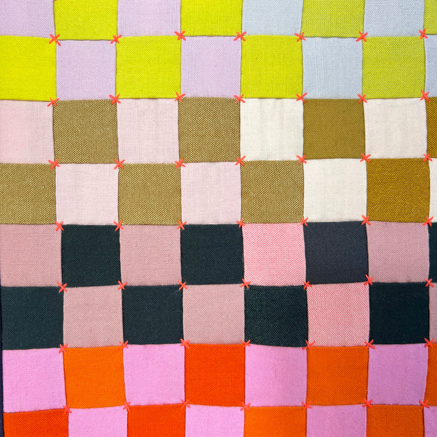 A close up of a small quilted wall art composed of multi colored squares  that is hand pieced, hand embroidered and stretched on archival acid free canvas.