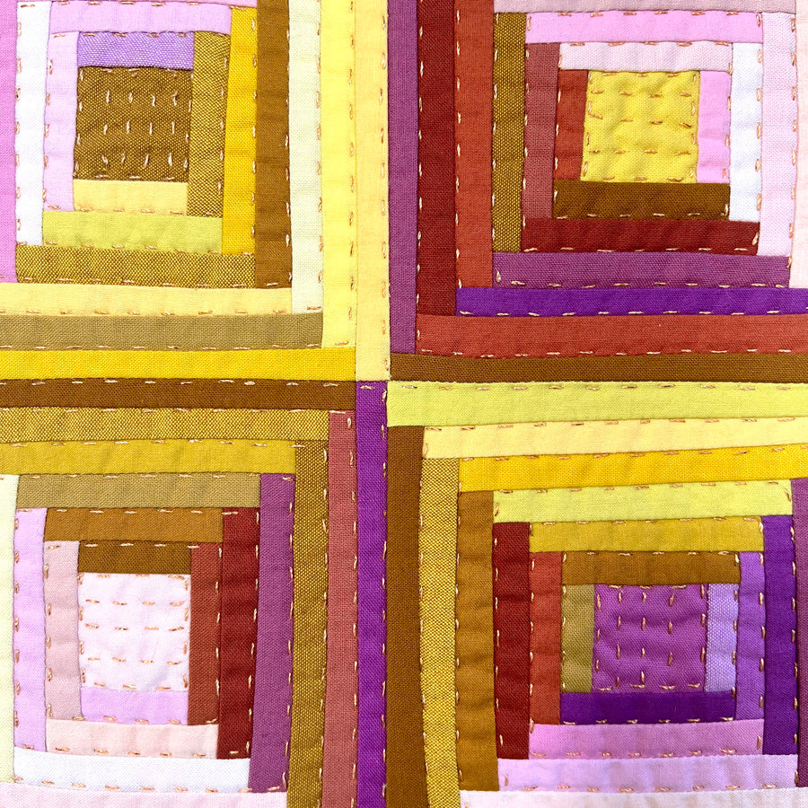 A close up of a small pieced log cabin block made of pink and yellow and brown fabrics, stretched on a canvas with metallic gold hand quilting.