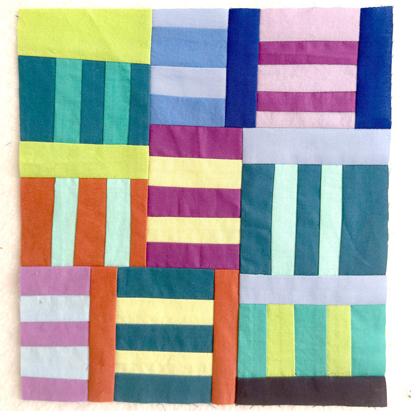 A square patchwork quilt or fabric panel made of multiple colorful stitched blocks with vertical and horizontal stripe patterns in yellow, teal, blue, pink, purple, orange, and light gray, photographed flat against a light neutral background.
