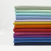 A stack of folded fabric pieces in a rainbow of colors, including mint green, light blue, lavender, yellow, orange, red, magenta, purple, blue, and navy, photographed on a clean white background.