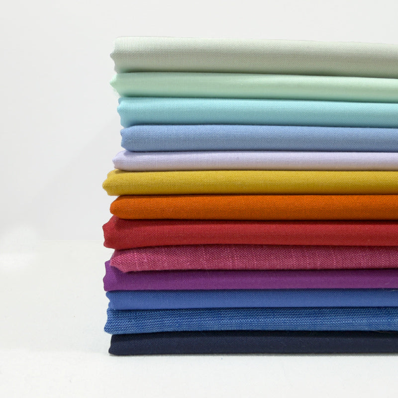 A stack of folded fabric pieces in a rainbow of colors, including mint green, light blue, lavender, yellow, orange, red, magenta, purple, blue, and navy, photographed on a clean white background.