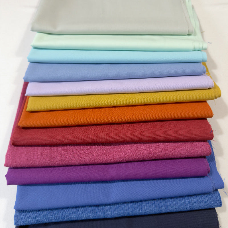 A stack of folded fabric pieces in a rainbow of colors, including mint green, light blue, lavender, yellow, orange, red, magenta, purple, blue, and navy, photographed on a clean white background.