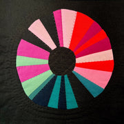 A wonky sewn circle on a black background. The circle is composed of wedges in shades of pink, blue and orange.