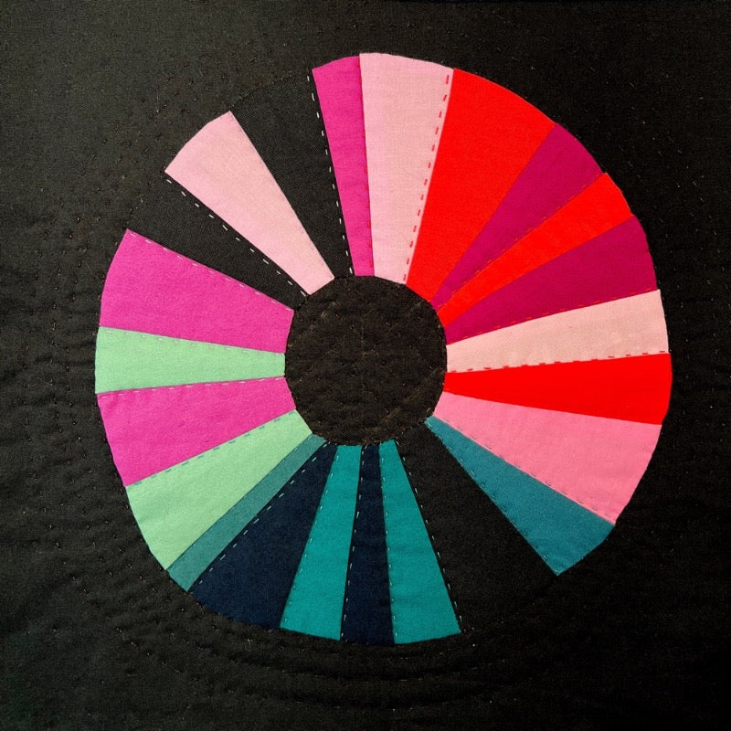 A wonky sewn circle on a black background. The circle is composed of wedges in shades of pink, blue and orange.