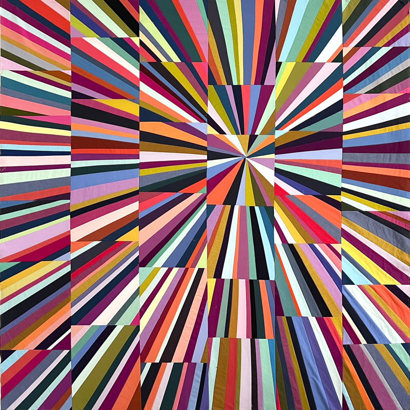 A square quilt with a vibrant, multicolored geometric pattern featuring radiating stripes and blocks of color from the center outward.