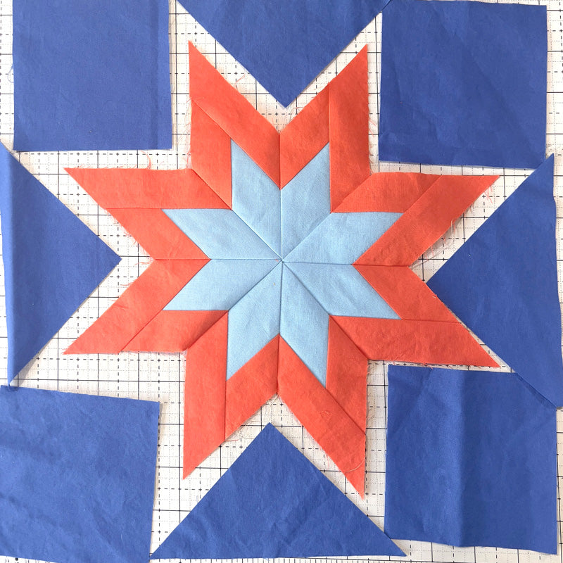 A quilt block featuring a geometric star pattern with a light blue center, orange-red star points, and blue corner blocks on a white gridded fabric background.